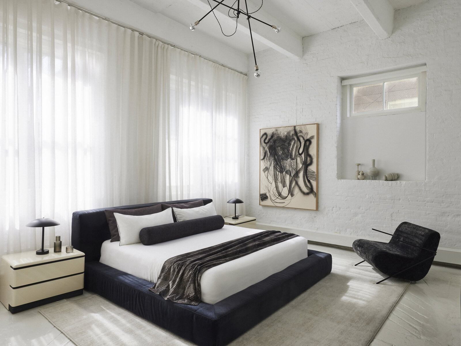 A white-brick loft bedroom with a navy platform bed, sheer curtains and a black gestural artwork, designed by JL Powers Design.