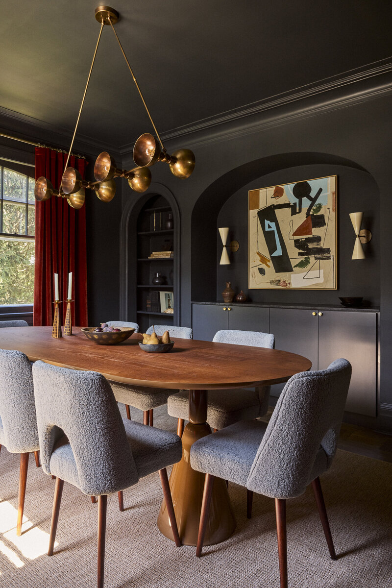 A charcoal dining room with a brass chandelier, oval walnut table, boucl&eacute; chairs and a red velvet curtain, designed by JL Powers Design.