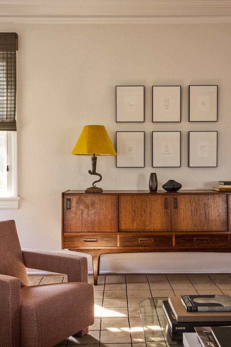 A living room with a rosewood credenza, yellow-shaded lamp and a grid of framed works on paper, designed by JL Powers Design.