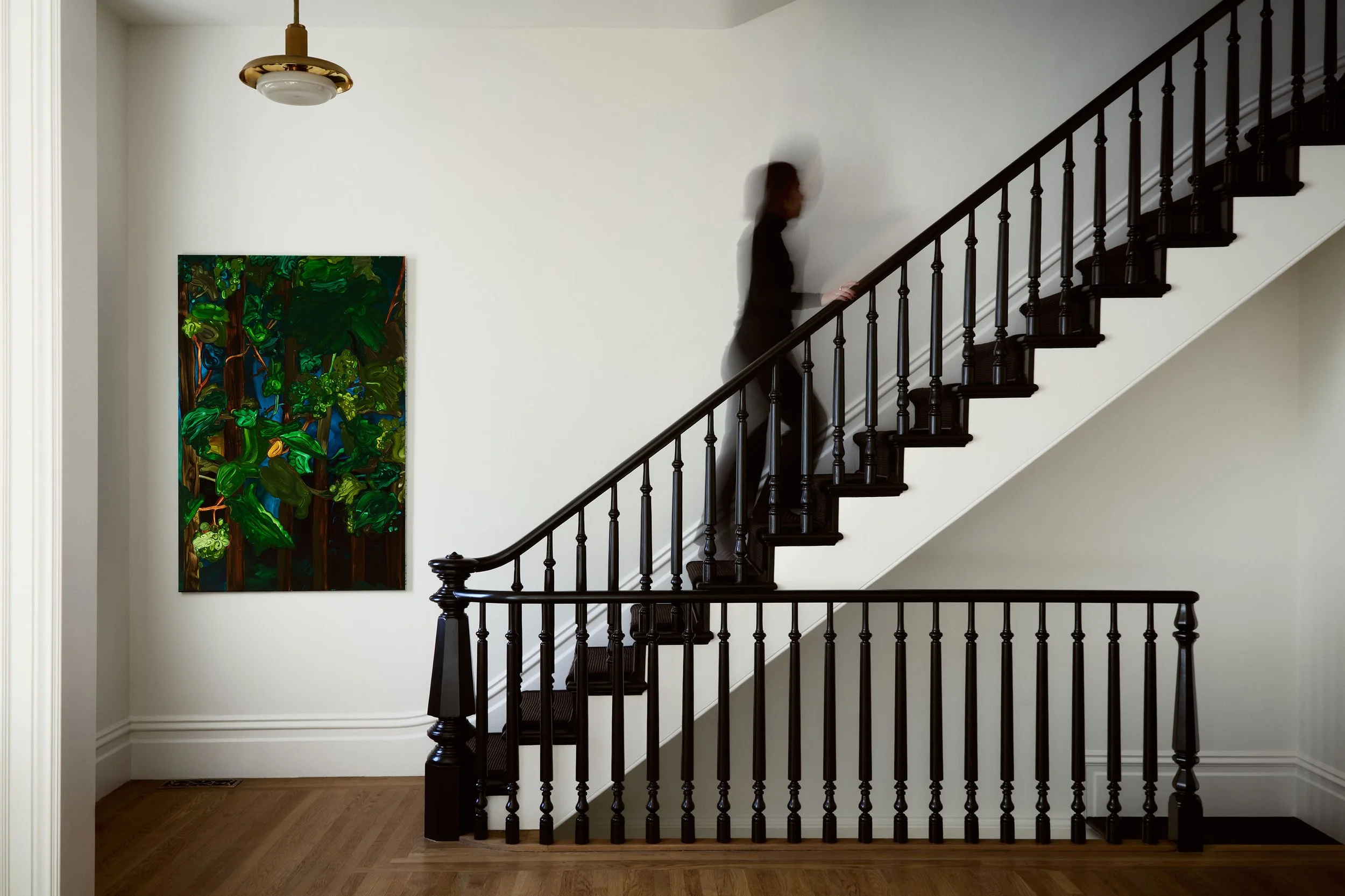 A designer walking up a dark-stained staircase in a white entry hall, a green forest painting on the wall and a brass pendant overhead.